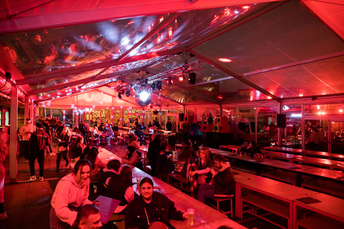 A marquee with red lighting and long bench seating with customers enjoying drinks