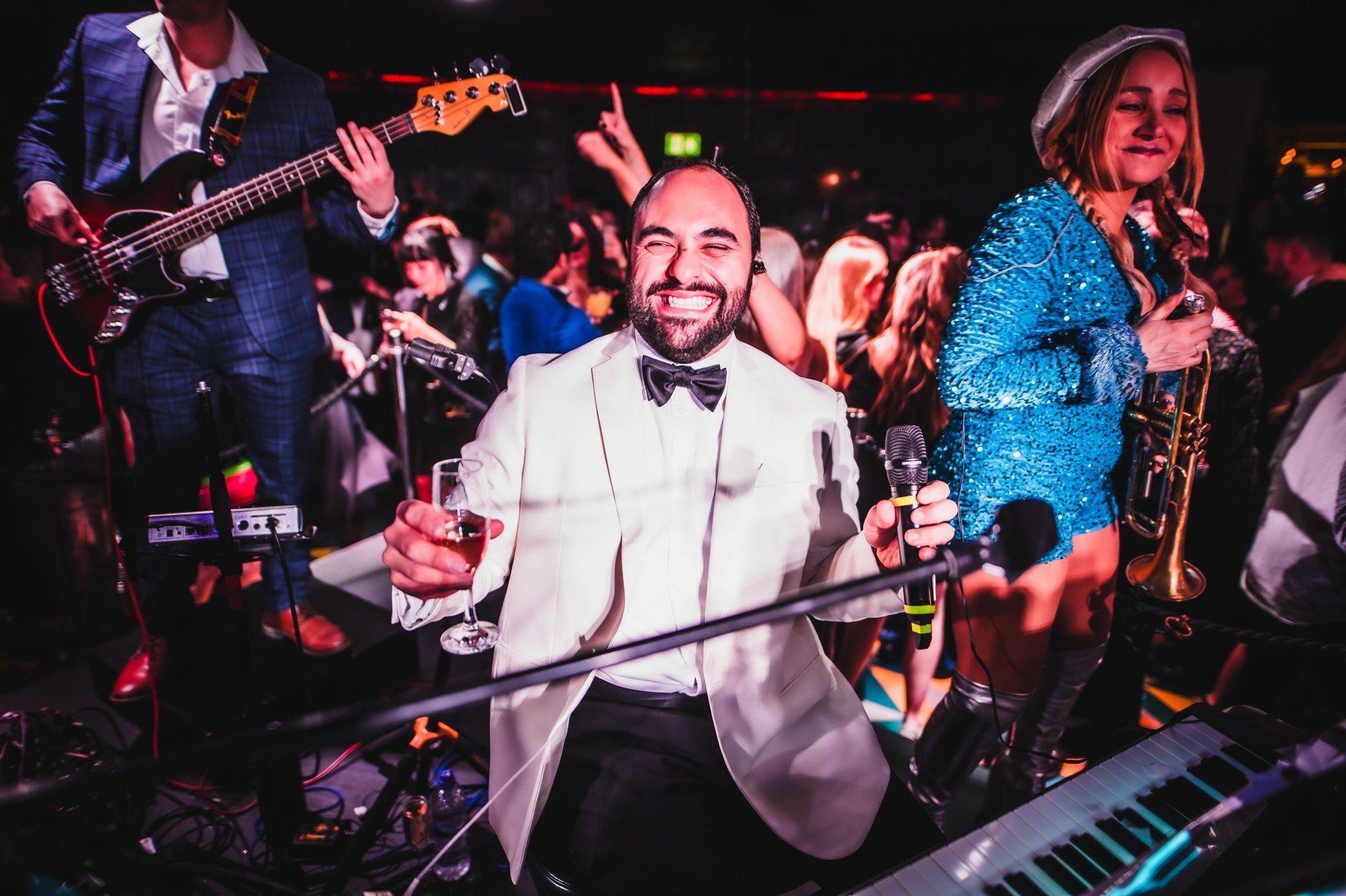 A man in a white tux holding a microphone and a glass of prosecco