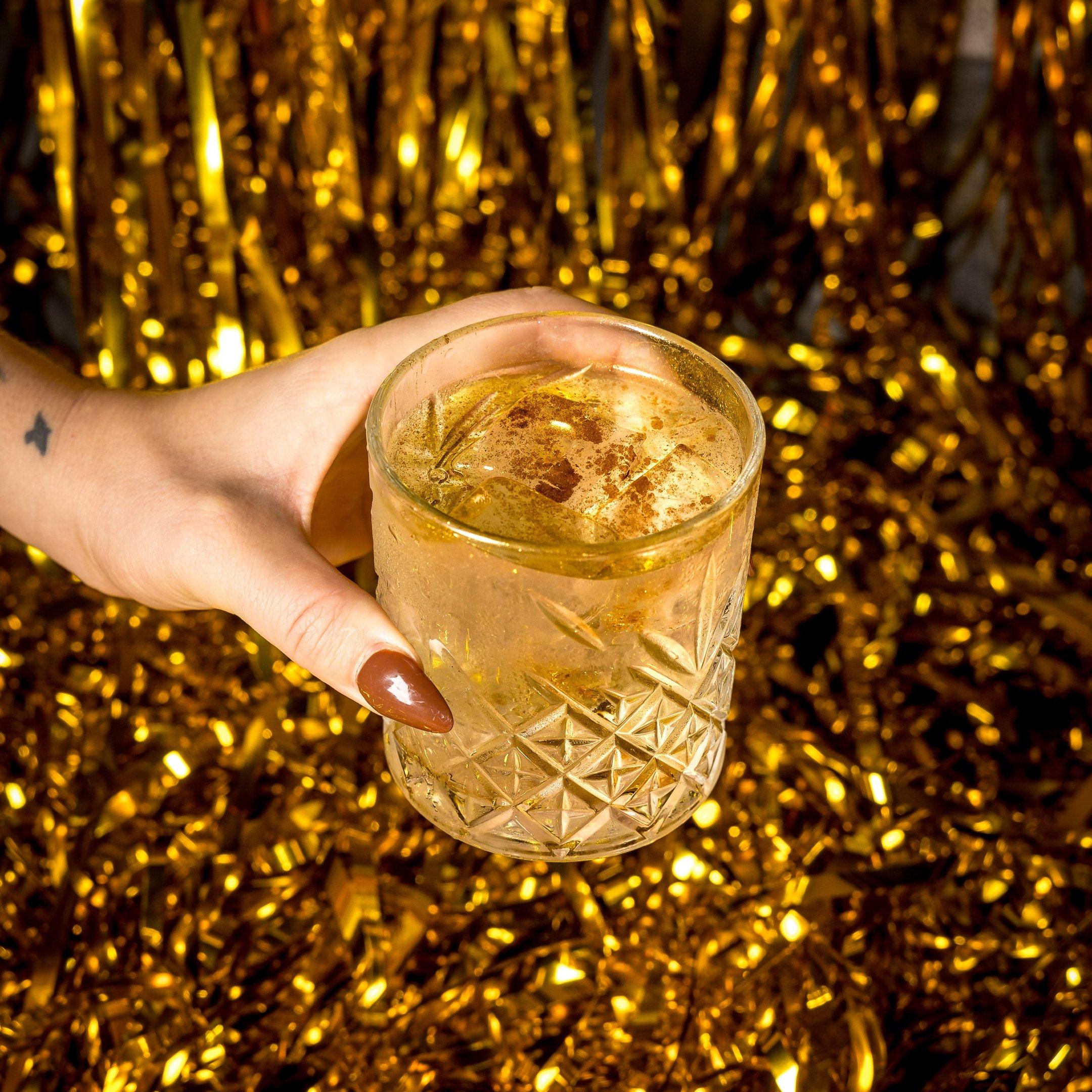 A hand holding an Unholy Water cocktail in front of a gold foil background