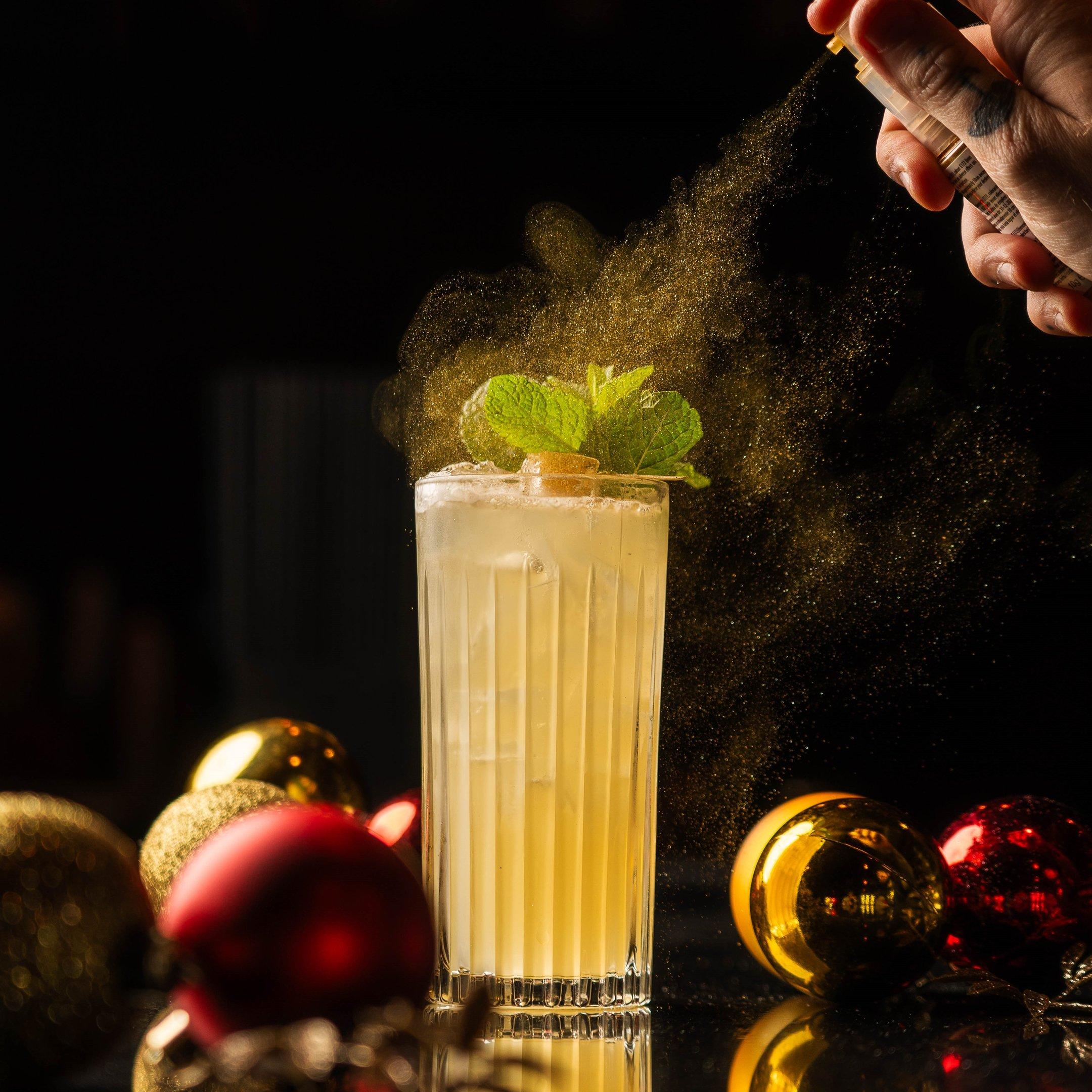 A cocktail in a tall glass being sprayed with a gold edible glitter