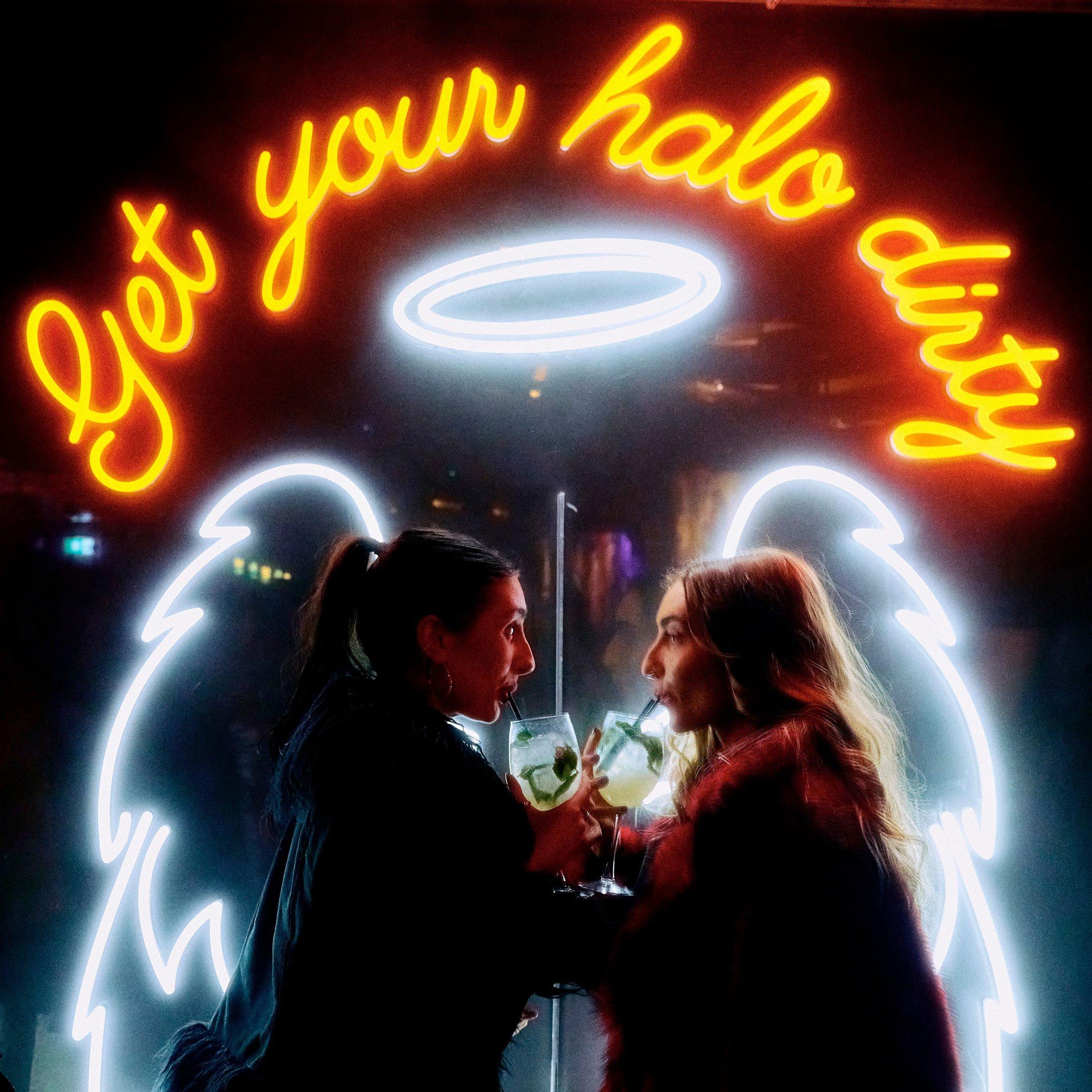 Girls sipping cocktails in front of an angel wings neon sign in Dirty Martini