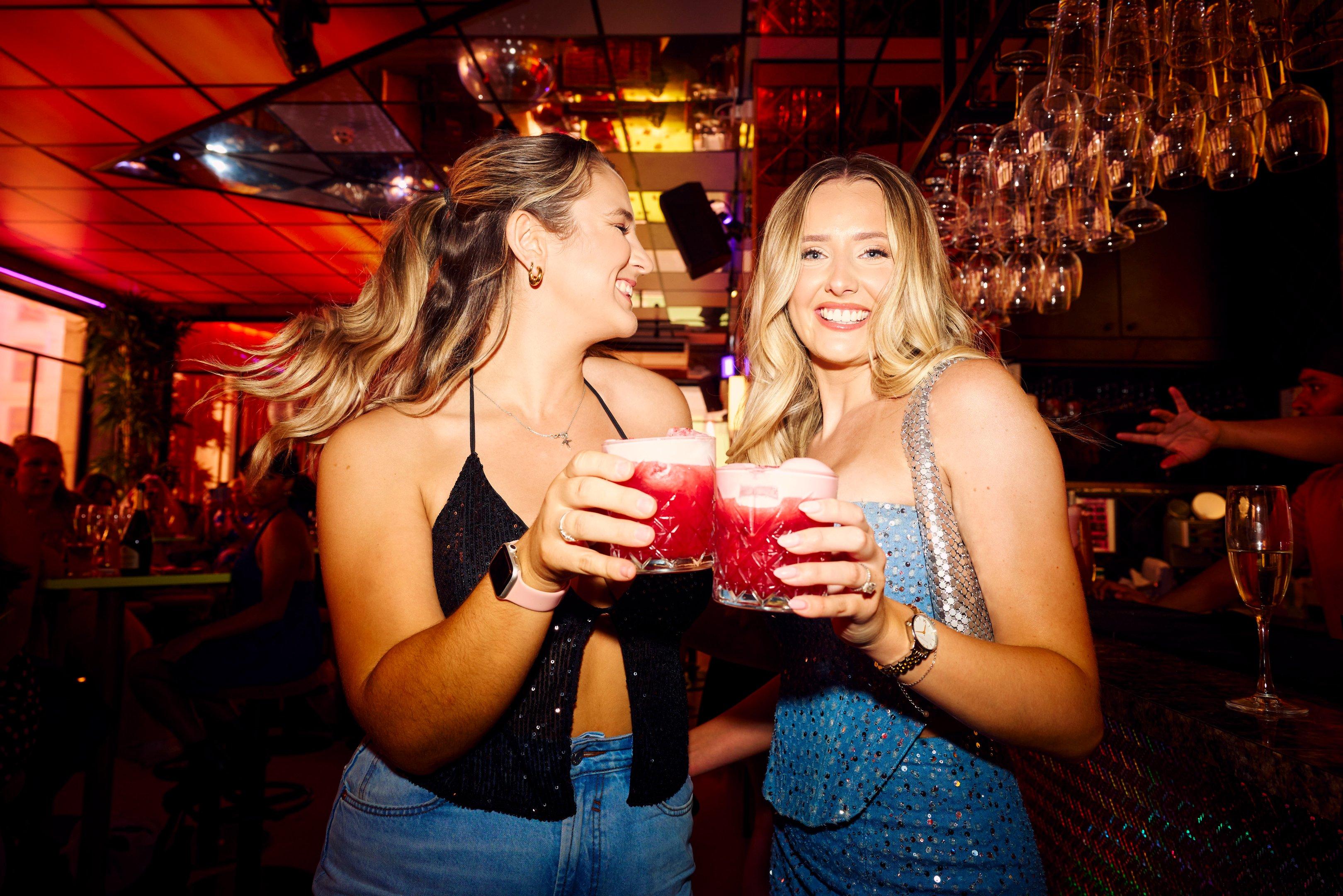 Two girls with cocktails in hand posing together for a picture in Nikki's bar