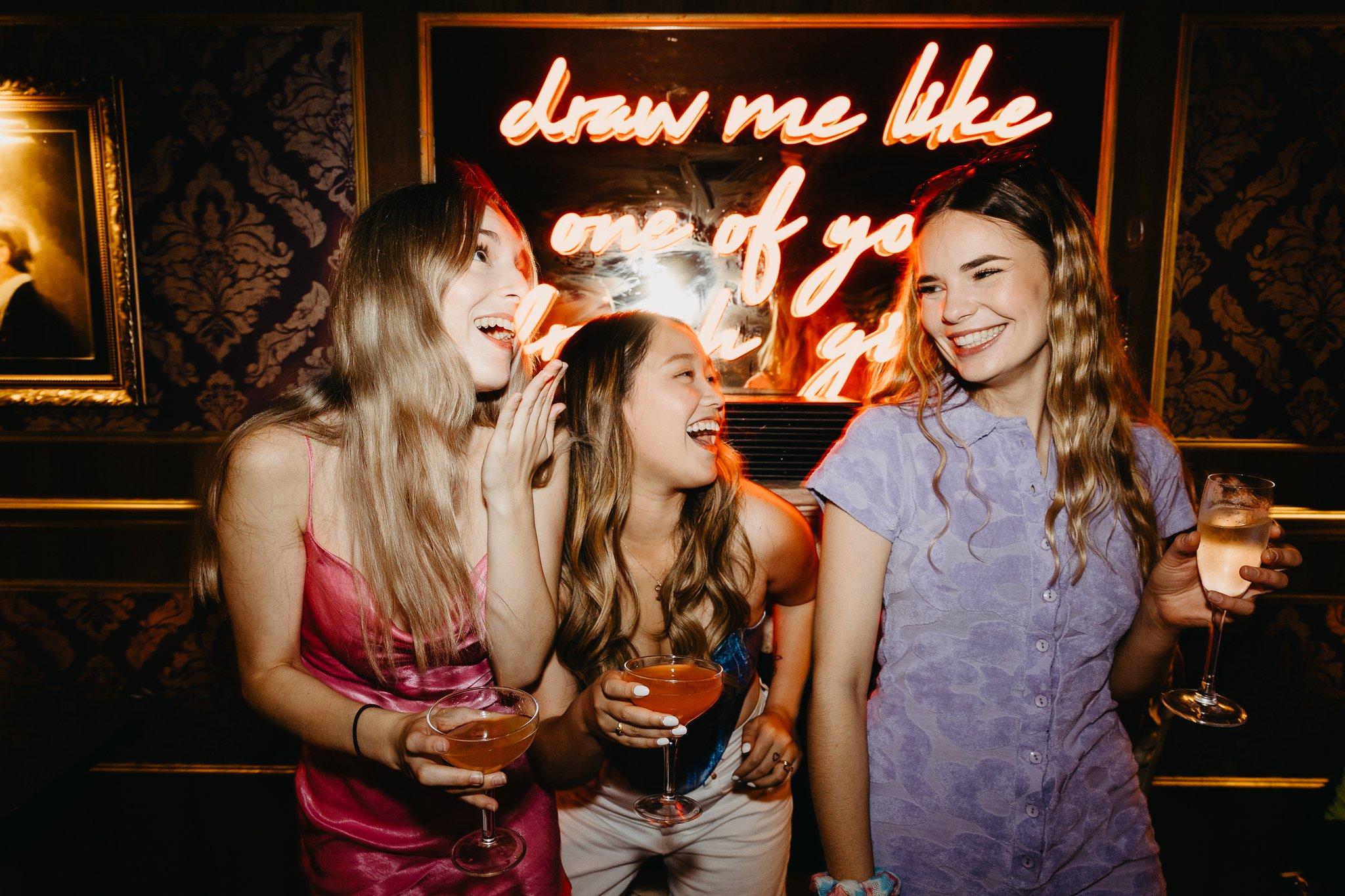 Three friends laughing with cocktails under a neon sign at The Cocktail Club