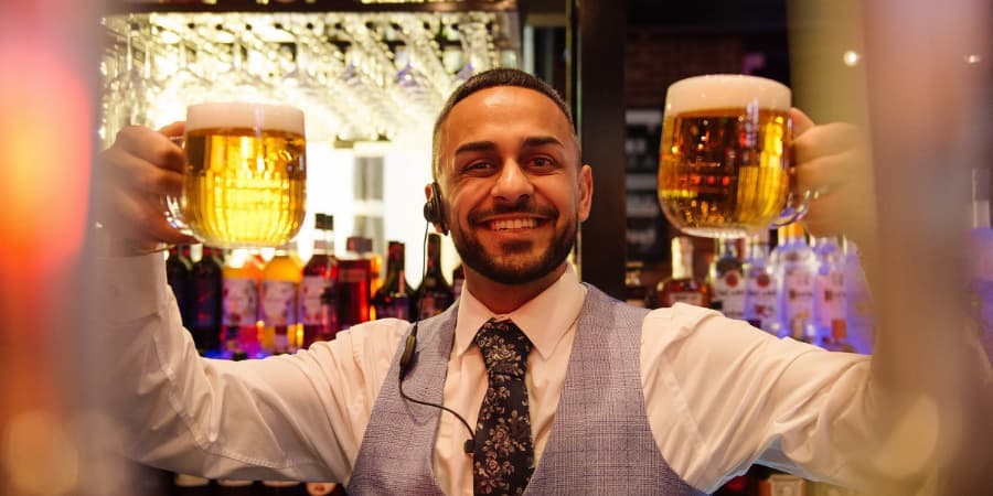 Barman holding two pints and smiling at the camera.