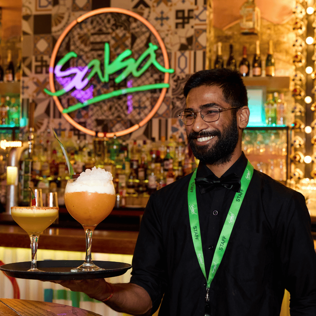 Bartender holding two cocktails on a tray in front of a Salsa! neon sign.