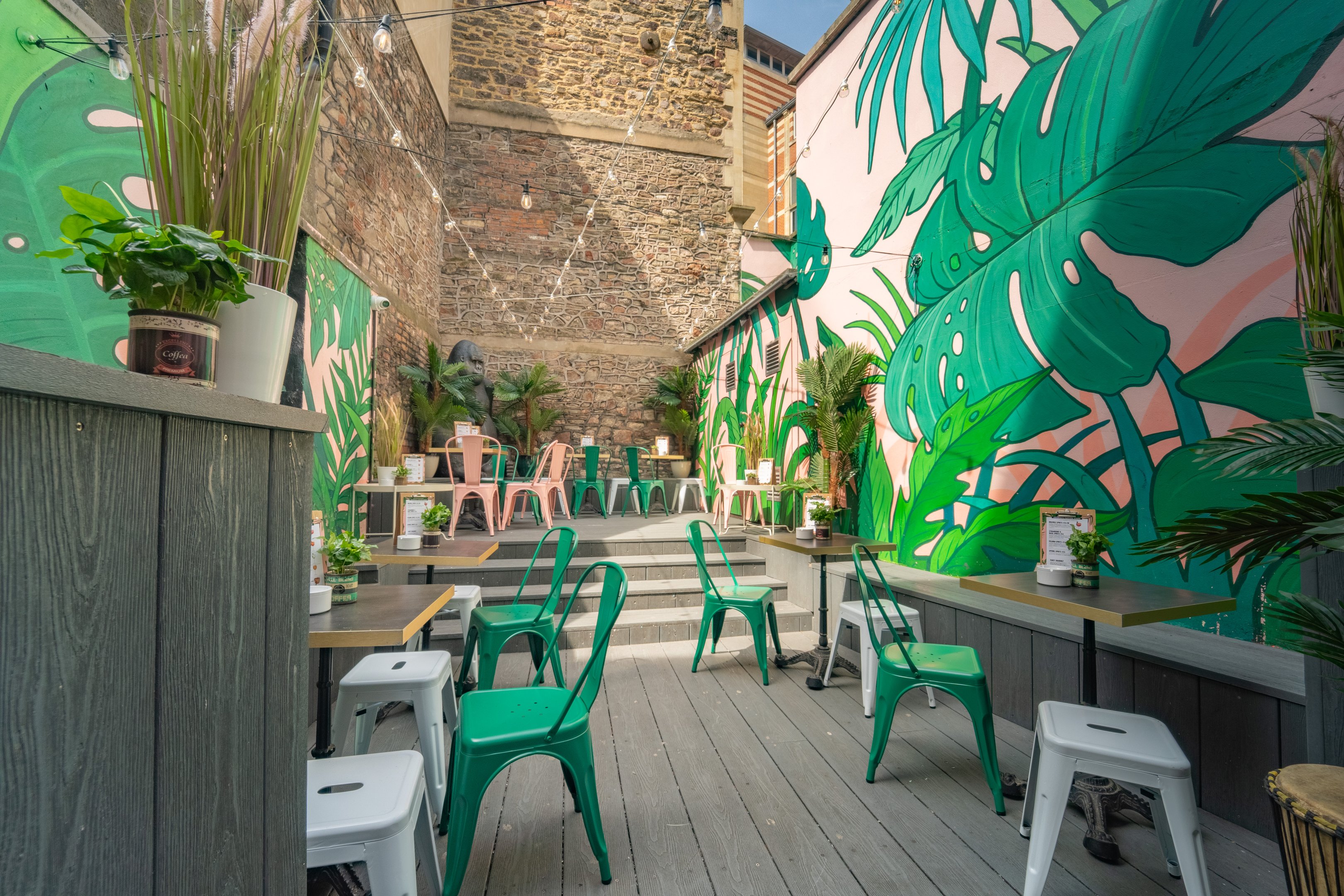 A shot of the Summer outdoor terrace with bold plant patterns on the walls and green and pink metal chairs and tables