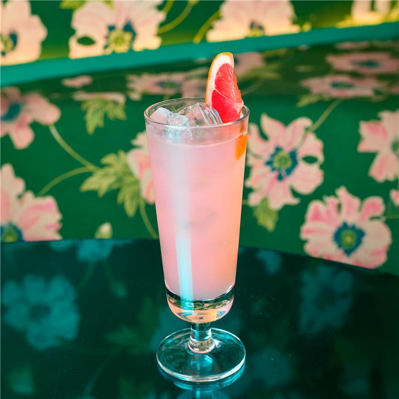 An image of a Paloma cocktail in a tall glass with a Grapefruit garnish sitting on a table in Blame Gloria